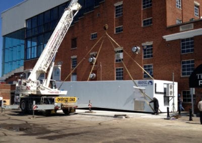 A white crane reaching the top of a brick building with a commercial freezer unit below